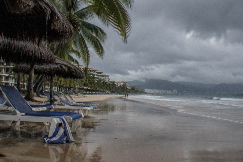 Empty beach chairs under palm trees beneath heavy gray monsoon clouds over the Pacific