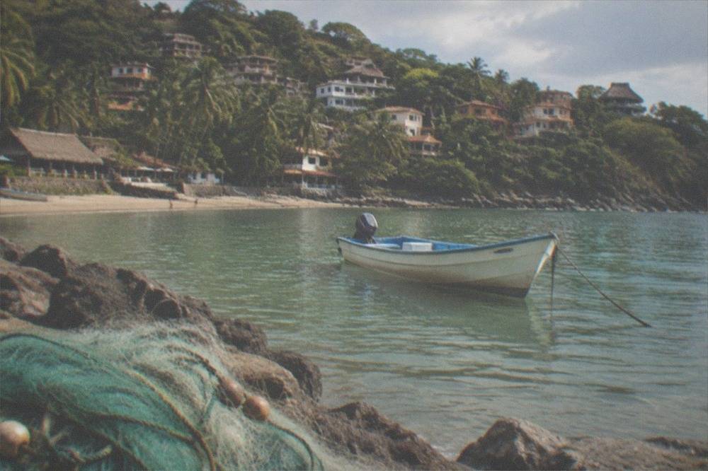 A small boat anchored in shallow water at a jungle-backed cove with a village on the hillside