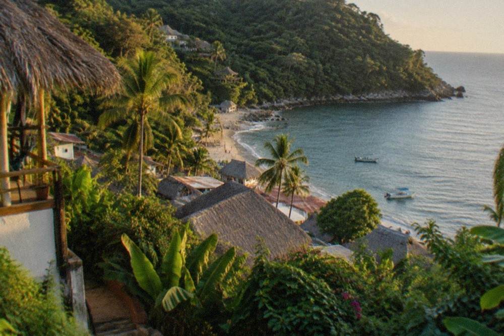 A jungle-covered hillside sloping down to a small beach town with palapa roofs along the shoreline