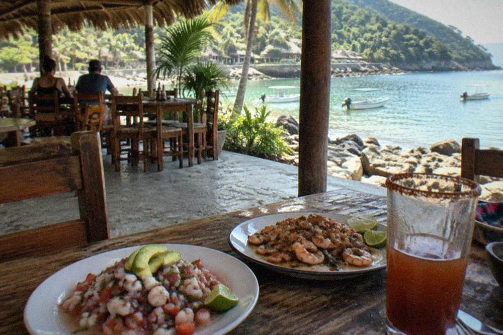 Open-air beachside restaurant with thatched roof and plates of seafood on wooden tables