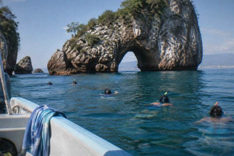 A panga idles beside the sea arches of Los Arcos in Banderas Bay, snorkelers floating in clear water close to the rocks.