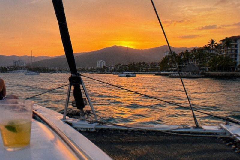 A catamaran glides across Banderas Bay at sunset, orange sky over the Sierra Madre and the Puerto Vallarta coastline beyond.