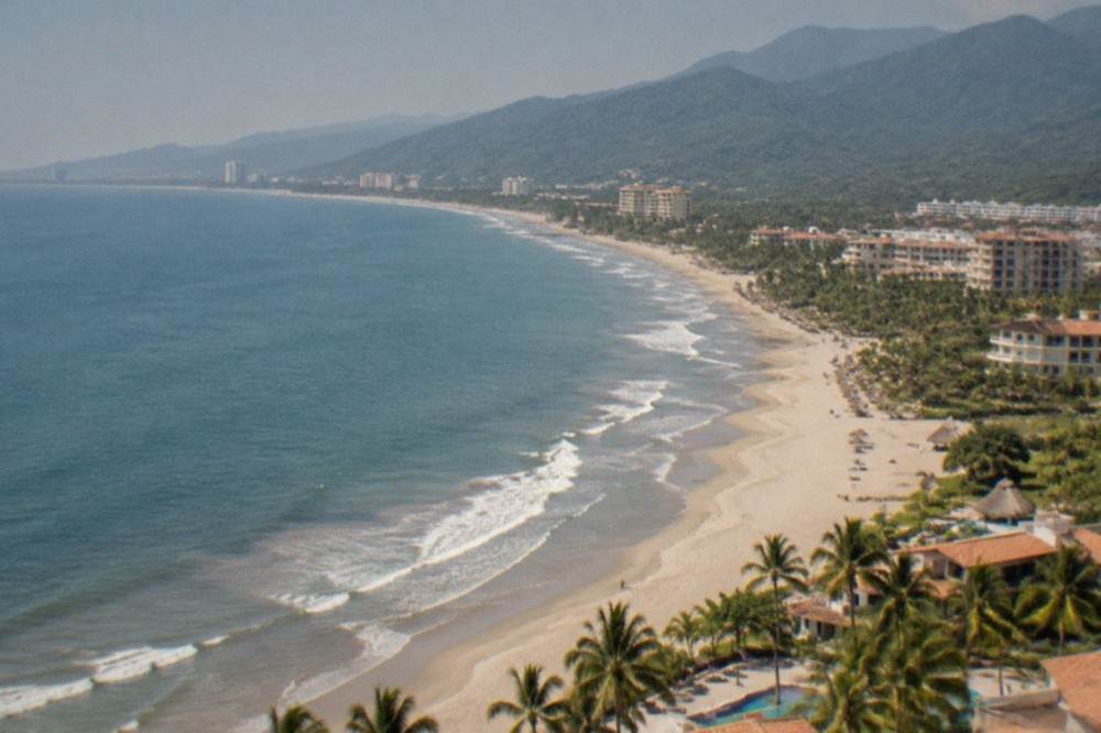 Wide aerial view of a large bay with blue water, a long sandy beach and dense green mountains at the edge