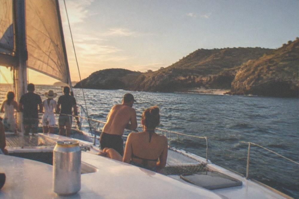 Catamaran with passengers on the upper deck passing a rocky coastline in late afternoon light